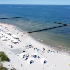 Majówka nad morzem: piaszczysta plaża z białymi parasolami, leżakami i budkami plażowymi, ludzie kąpią się i opalają.