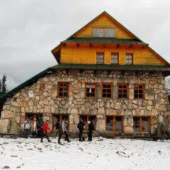Kamienny murowaniec wysokosc w górach, otoczony śniegiem i lasem. Ludzie w zimowych ubraniach zbliżają się do schroniska.