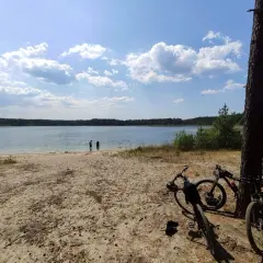 Dwa rowery czekają na plaży nad jeziorem, gotowe na odkrywanie **Swornegacie trasy rowerowe**. W tle woda i las.