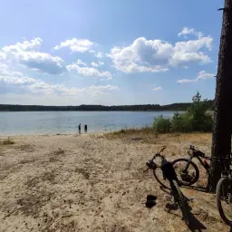 Dwa rowery czekają na plaży nad jeziorem, gotowe na odkrywanie **Swornegacie trasy rowerowe**. W tle woda i las.