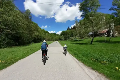 Dwie osoby na rowerach jadą drogą przez zieloną dolinę. W tle widać dom i skały. Idealny dzień na wavelo!