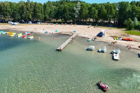 Słoneczny dzień nad jeziorem, gdzie kolorowe rowerki wodne i kajaki czekają na plaży. W tle widać namioty i kampery, tworząc atmosferę letniego binduga camping.