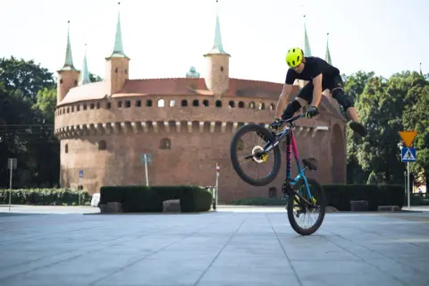 Wykonuje trik na rowerze po krakowsku, z zabytkowym Barbakanem w tle.