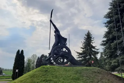 Pomnik Kościuszki w Racławicach, z kosynierem na armacie, pod pochmurnym niebem.