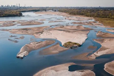 Widok z lotu ptaka na rozległe piaszczyste łachy i meandrujące koryta rzeki, ukazujący niski stan Wisły. W tle majaczy panorama miasta.