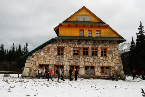 Kamienny murowaniec wysokosc w górach, otoczony śniegiem i lasem. Ludzie w zimowych ubraniach zbliżają się do schroniska.