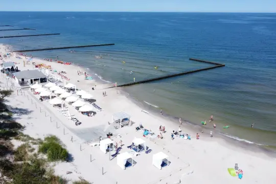 Majówka nad morzem: piaszczysta plaża z białymi parasolami, leżakami i budkami plażowymi, ludzie kąpią się i opalają.