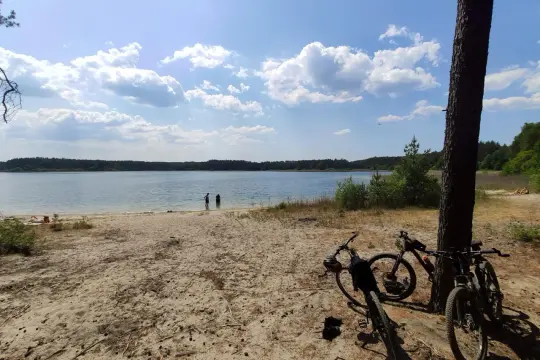 Dwa rowery czekają na plaży nad jeziorem, gotowe na odkrywanie **Swornegacie trasy rowerowe**. W tle woda i las.