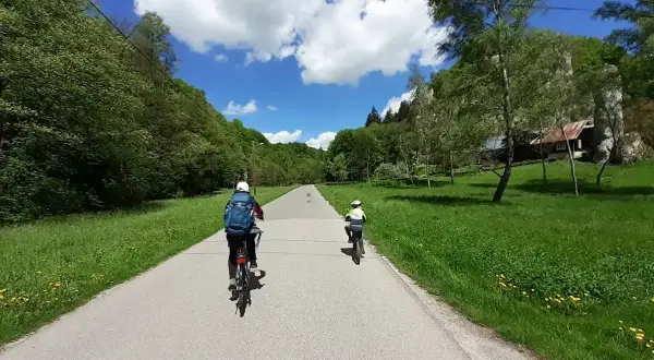 Dwie osoby na rowerach jadą drogą przez zieloną dolinę. W tle widać dom i skały. Idealny dzień na wavelo!
