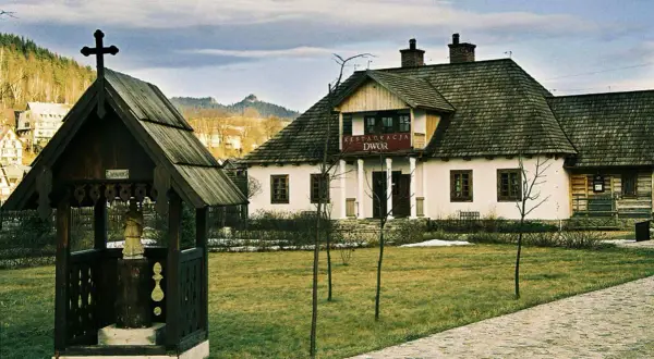Drewniana kapliczka z krzyżem i figurą, obok restauracja "Dwór". W tle sosny i zabudowania. Idealne miejsce na camping.