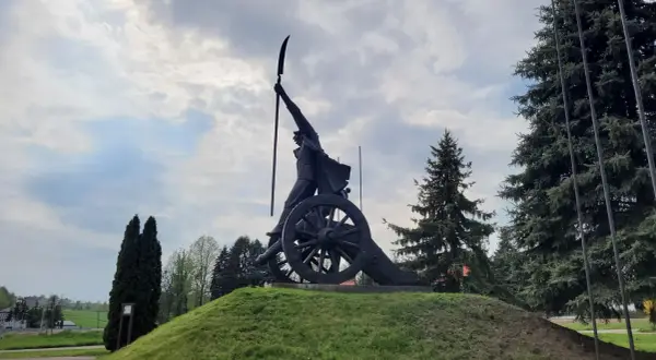 Pomnik Kościuszki w Racławicach, z kosynierem na armacie, pod pochmurnym niebem.
