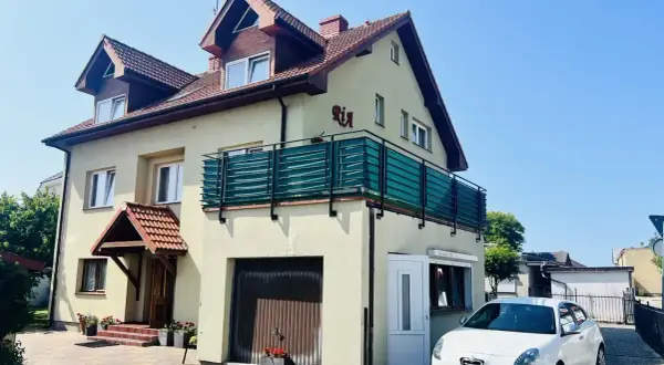 Jasny dom z balkonem i napisem "Ria" oferuje kwatery nad morzem. Biały samochód zaparkowany przed posesją.