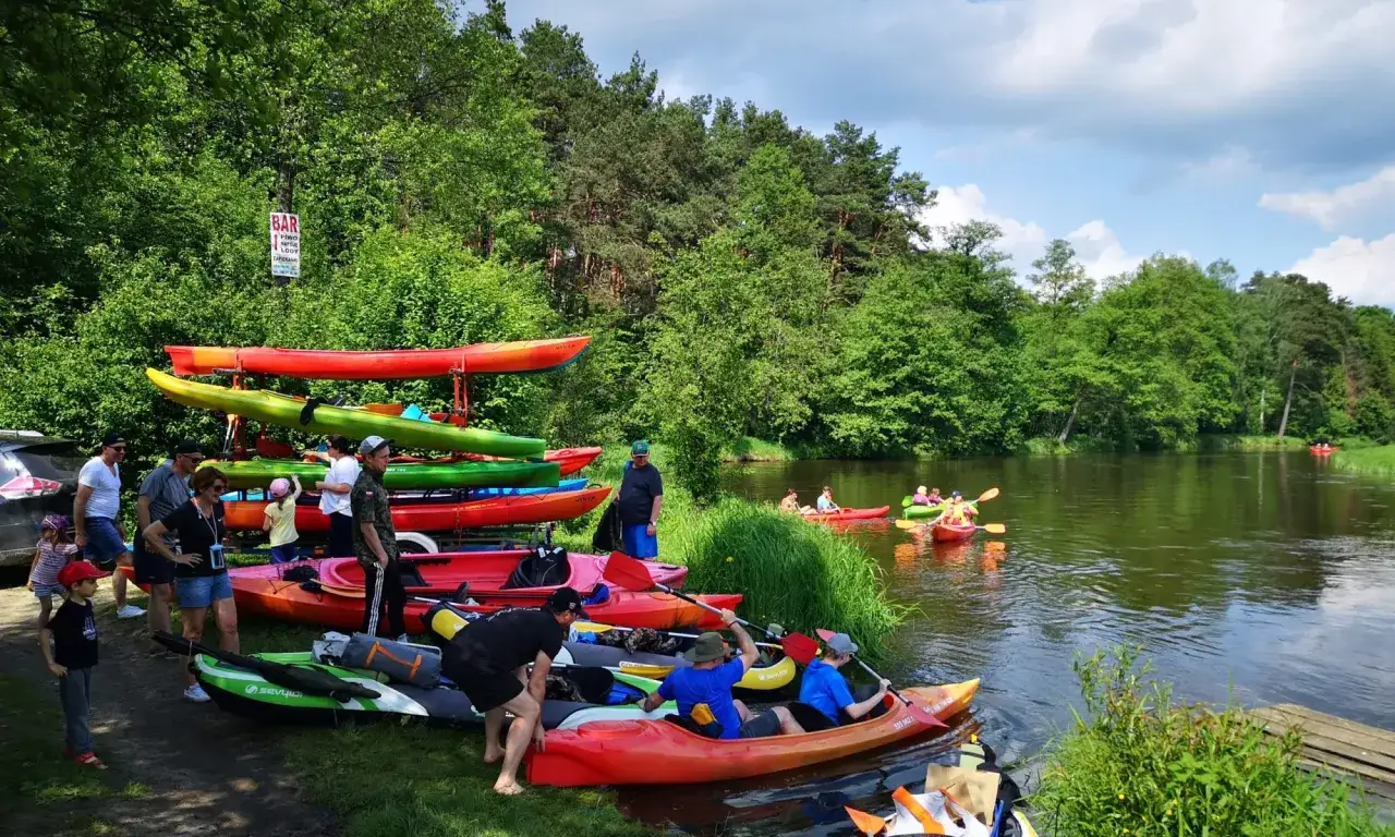 Ludzie przygotowują się do spływu kajakowego po Pilicy. Widać wiele kajaków na brzegu i kilka już na wodzie. Pilica głębokość jest wystarczająca do bezpiecznego pływania.