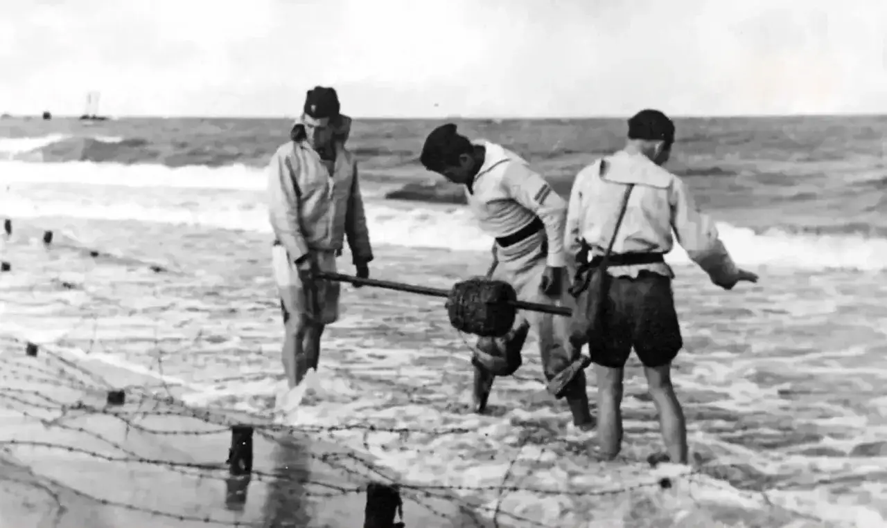 Żołnierze pracują przy budowie fortyfikacji, tworząc zaporę z drutu kolczastego na plaży. W tle widać morze i statki, sugerując strategiczne znaczenie tych bunkrów nad morzem.