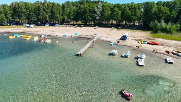 Słoneczny dzień nad jeziorem, gdzie kolorowe rowerki wodne i kajaki czekają na plaży. W tle widać namioty i kampery, tworząc atmosferę letniego binduga camping.
